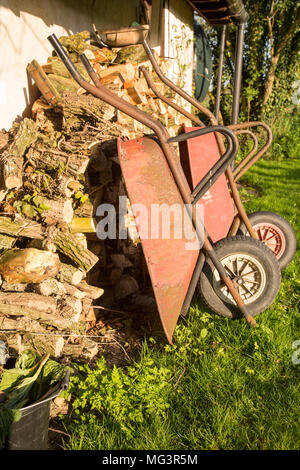 Two old red metal wheelbarrows leaning against pile wood logs in garden, Cherhill, Wiltshire, England, UK Stock Photo