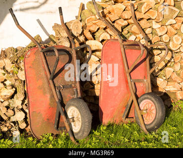 Two old red metal wheelbarrows leaning against pile wood logs in garden, Cherhill, Wiltshire, England, UK Stock Photo