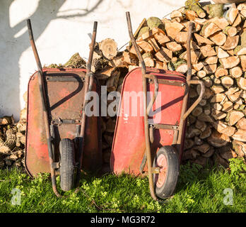 Two old red metal wheelbarrows leaning against pile wood logs in garden, Cherhill, Wiltshire, England, UK Stock Photo
