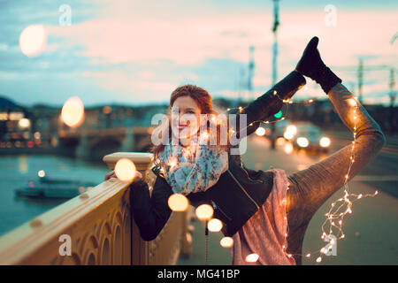 Happy young redhead woman doing yoga with fairy lights outdoors and smile, teal and orange style Stock Photo