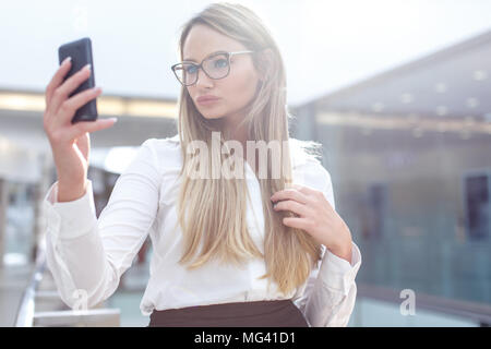 Young smart blonde businesswoman in eyeglasses taking selfie in business center, indoors Stock Photo