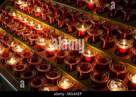 Small white votive candles lighted and placed in red glass candlesticks among empty ones, lined up on a candle holder stand in a catholic church. Stock Photo