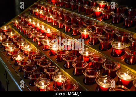 Small white votive candles lighted and placed in red glass candlesticks among empty ones, lined up on a candle holder stand in a catholic church. Stock Photo