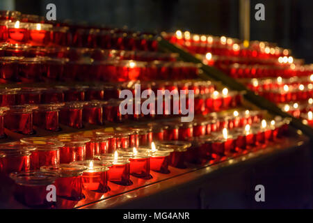 Small white votive candles lighted and placed in red glass candlesticks among empty ones, lined up on a candle holder stand in a catholic church. Stock Photo