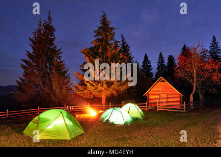 Night camping. Tourist have a rest at a campfire near illuminated tent and wooden house under amazing night sky full of stars and milky way. Astrophotography Stock Photo