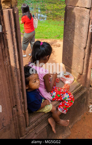 Two local kids, a little boy and a young girl, are sitting barefoot on a window frame ruin in the Banteay Srei (Citadel of the Women) temple. Stock Photo