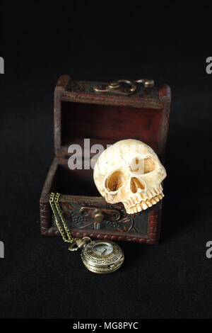 an evil human skull with a pocket watch on a pirate treasure chest on a black background Stock Photo