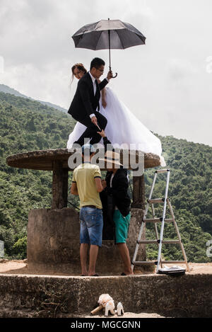 Newly-wed couple, posing for photographs, perched on an old well, holding umbrella, while photographer and assistant talk below, Hue, Vietnam Stock Photo