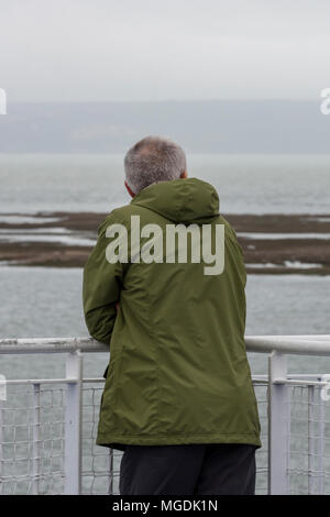 A man wearing a green outdoor coat leaning on somew railings looking out to sea over the beaches and marshlands on the lymington river in Hampshire uk Stock Photo