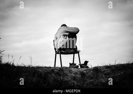 A poor refugee refugee in the Jungle field of Calais crouches hooded on a chair at the top of the hill taking off his shoes Stock Photo