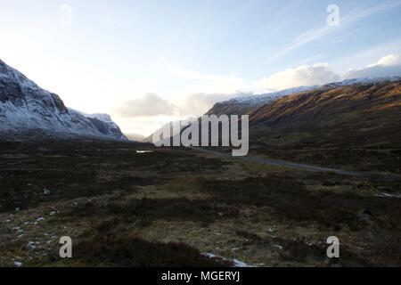 A concrete road crosses the Scottish Highlands at sunset with snowy peaks and typical autumn colors at the bottom of the valley Stock Photo