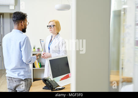 Pretty blond-haired physician wearing white coat and eyeglasses standing at spacious office while consulting bearded patient Stock Photo