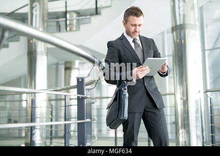 Serious busy handsome manager using digital tablet to have access to files: he standing in office corridor and holding briefcase Stock Photo
