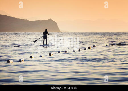Silhouette of a man paddling on a surfboard towards the open sea alongside the floats at sunset time Stock Photo