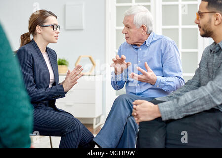 Aged patient wearing denim shirt having productive conversation with highly professional psychologist while participating in group therapy session at  Stock Photo