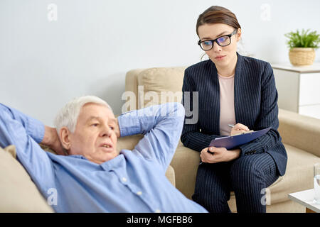 Depressed senior man lying on cozy sofa of psychotherapy office and talking to highly professional young psychologist, she taking necessary notes Stock Photo