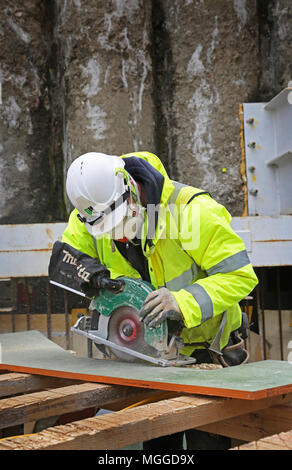 A workman (unidentifiable) uses an electrical circular saw to cut plywood on a London construction site. Stock Photo