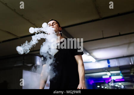 Man in cap smoke an electronic cigarette and releases clouds of vapor performing various kind of vaping tricks Stock Photo