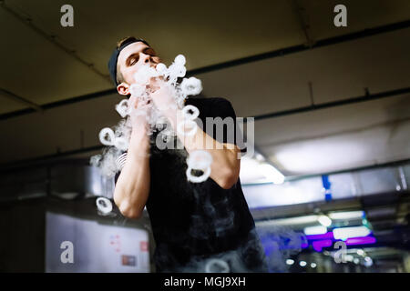Man in cap smoke an electronic cigarette and releases clouds of vapor performing various kind of vaping tricks Stock Photo
