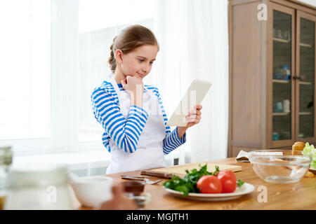 Curious little girl wrapped up in reading recipe with help of digital tablet while standing at wooden kitchen table in order to make surprise for Moth Stock Photo