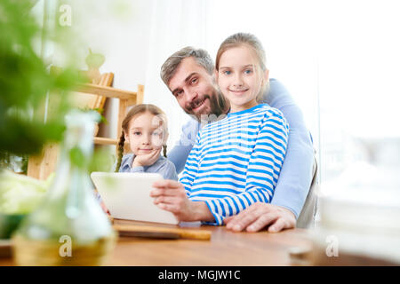 Joyful middle-aged father and his adorable little daughters posing for photography while using digital tablet in order to write down menu items for bi Stock Photo