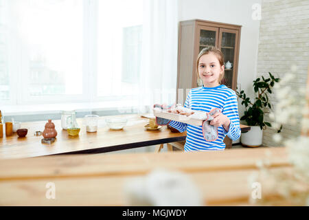 Portrait shot of cute little girl holding baking tray with just prepared cupcakes in hands and looking at camera while making surprise for Mothers Day Stock Photo