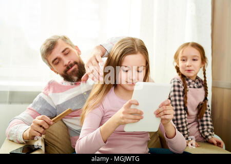 Loving dad spending weekend at home: he combing hair of little daughter while she browsing Internet on digital tablet, her cute sister keeping eye on  Stock Photo