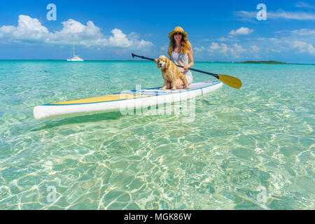 The Bahamas - a blonde woman in a hat sits on a paddleboard with her  cute, tan cocker spaniel. Sailboat in background on clear green water at a sandy Stock Photo