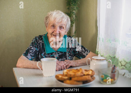 An elderly woman drinking tea in the kitchen. Looks into the camera. Stock Photo