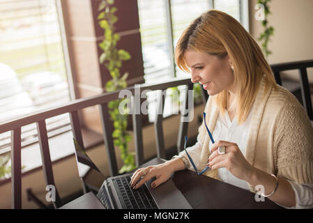 Blonde business woman is working on laptop in coffee shop, restaurant. Charming happy women using computer to prepare for the course work. Looking for Stock Photo