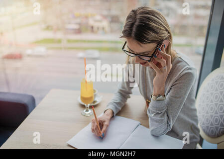 Business woman talking on smartphone, taking notes in a coffee shop, restaurant. Gorgeous woman taking a notes to prepare for the course work. Drinkin Stock Photo