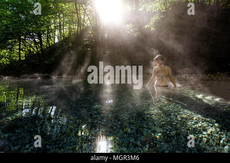 Young woman in bikini standing in a natural hot pool in the forest with backlight and steam around her, Maibachl, Austria. Stock Photo