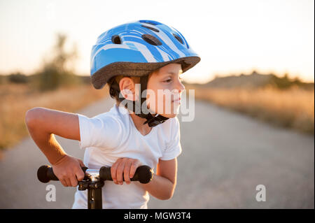 adorable little kid in sport helmet standing on abandoned road with scooter in warm summer sunset Stock Photo