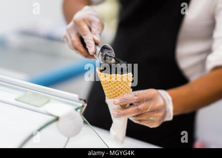 Handsome young waiter in elegant classic suit putting Black ice cream balls in waffle cone. Trendy dessert. Real scene. Professional service, catering Stock Photo