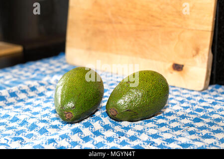 Two whole avocados on a blue and white towel with a wooden chopping board in the background Stock Photo