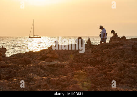 Two tourists walking at sunset and a sailing ship in the background at Cap de Ses Salines cape in Majorca (Balearic Islands, Mediterranean Sea, Spain) Stock Photo