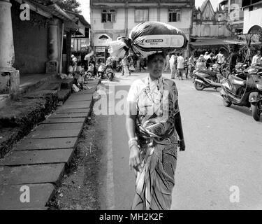 A woman carrying a large, heavy bag of rice balanced on her head through the street in Margao, near Benaulim, Goa, India Stock Photo