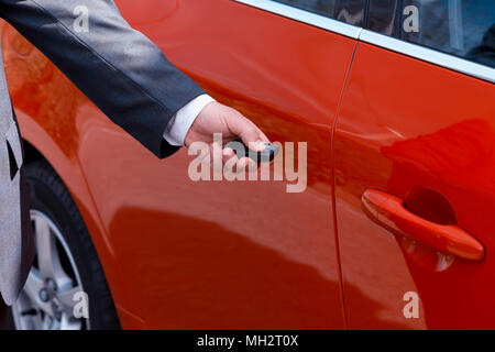 A man dressed in a gray suit opens or closes his modern red car. Stock Photo
