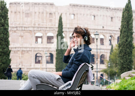 Handsome young man in sports cloths listening to music sitting on a bench with Colosseum in the background. Young sportsman in tracksuit relaxing afte Stock Photo