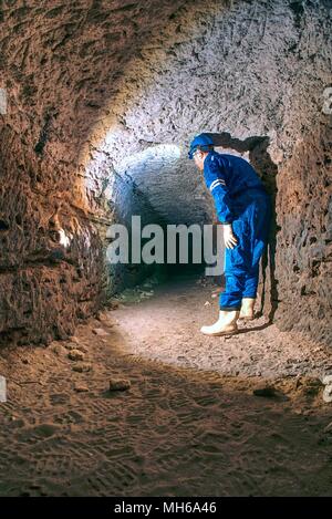 Dry sandstone tunnel, man worker in protective suite in underground. Mysterious dungeon tunnel with walls made inorange sandstone rock Stock Photo