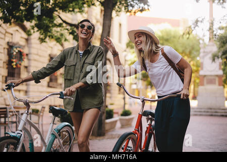 Positive and happy girls walking on the city street with bicycles. Female friends enjoying a walking down the street with their bikes. Stock Photo