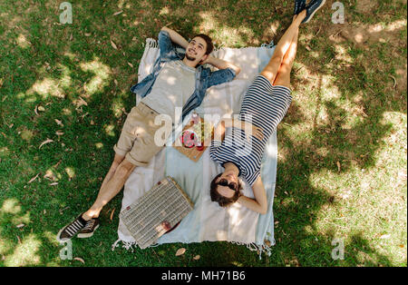 View from above of relaxed man and woman lying down at the park with hand basket, appetizing snacks and beers on blanket. Couple on picnic lying on bl Stock Photo