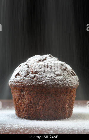 Close up of a delicious chocolate muffin on a wooden table, against black background, with snow like powder sugar scattered around and falling above.  Stock Photo