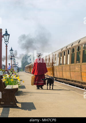 Odd lady, rear view, in costume (Mary Poppins) walking on platform at vintage railway station dog on lead, holding bag, looking at carriages to board. Stock Photo