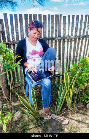 A beautiful Filipino woman sits in a chair at the beach using her iPad to stay in touch with friends in Palawan, Philippine Islands. Stock Photo