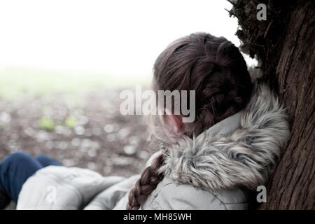 A young girl (aged eight), wrapped up warm in a fur-lined coat, leans against a tree with her back to the camera, in a slumped pose Stock Photo