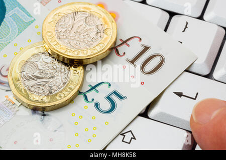 Sterling £5 and £10 pound notes with new pound coins pounds on a keyboard with a person's finger pressing enter key to buy online shopping concept. UK Stock Photo