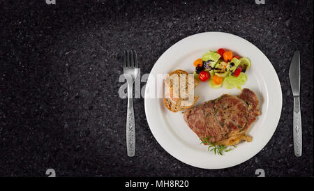 Ripened seasoned beef rump or striploin steak cooked on white plate with salad and home made bread. Top view on granite background with copy space. Stock Photo