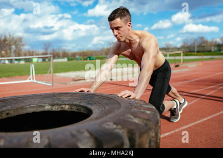 Young muscular man pushing up a huge tire for training muscles at the tartan track on the stadium. Stock Photo