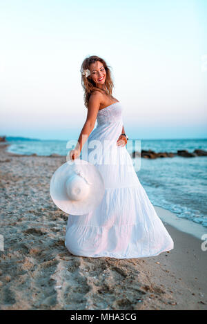 Beautiful young smiling woman in white dress walking on the beach and  holding white summer hat Stock Photo - Alamy
